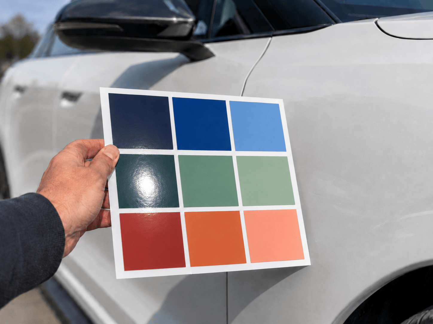 Hand holding a vinyl film sample against a vehicle in sunlight